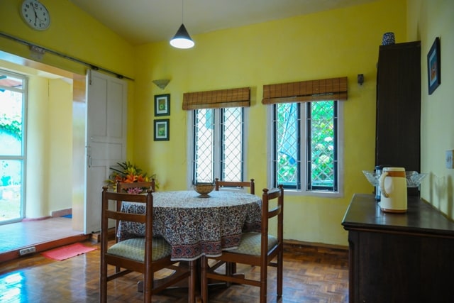 A brightly lit dining area features a round table surrounded by wooden chairs. The warm yellow walls complement the natural light streaming through the three windows, which are adorned with bamboo shades, providing a cheerful ambiance.