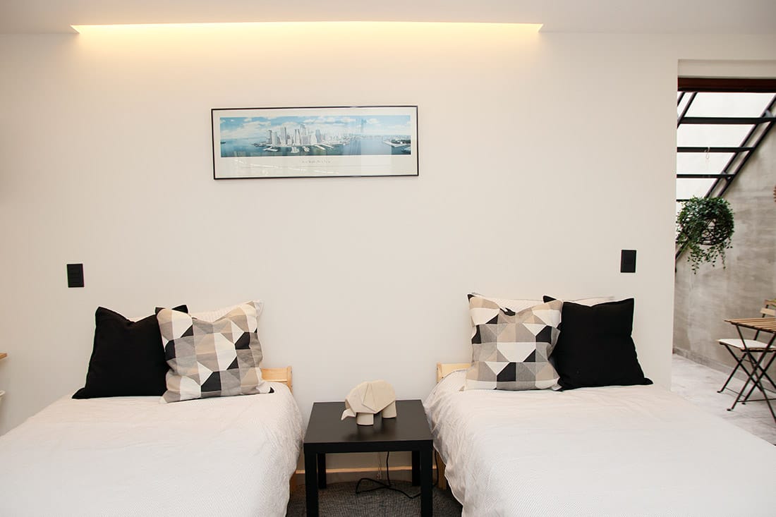 The sleeping area features two individual beds arranged side by side, both dressed in white linens. Each bed is accompanied by decorative pillows in geometric patterns. A small black table sits between the beds, and a framed artwork adorns the wall above.