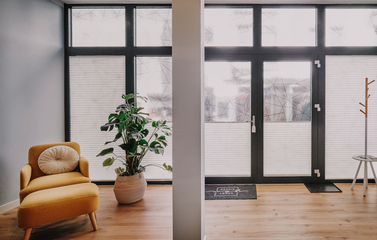 A bright entryway features large windows with patterned glass that allow natural light to filter in. A vibrant yellow chair is positioned beside a potted plant, while a small table stands near the door, enhancing the welcoming atmosphere of the space.