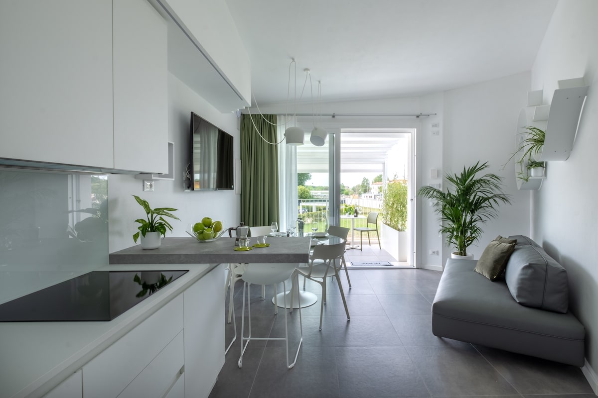 A modern kitchen and dining area features sleek white cabinetry and a gray countertop. A small dining table with white chairs is positioned near glass doors that open to a balcony, offering views of greenery. A comfortable gray sofa is located in the living space.