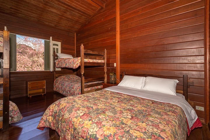 Drevená izba s manželskou posteľou a poschodovou posteľou, obe s kvetinovými posteľnými prikrývkami, ktoré prinášajú útulný a vidiecky nádych. Otvorené okno odhaľuje očarujúcu krajinu s kvetmi v pozadí, ktorá dotvára atmosféru útočiska v prírode.