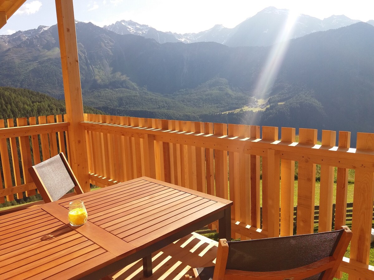 A wooden terrace is shown, featuring a dining table with a smooth surface and two comfortable chairs. The backdrop includes sweeping views of mountainous terrain, with sunlight casting a gentle beam across the landscape.