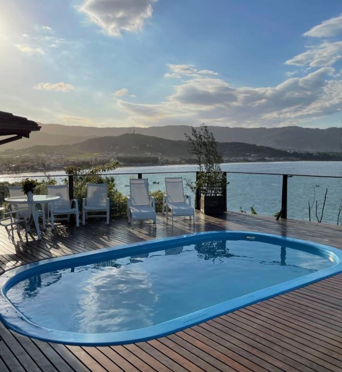 An outdoor deck features a heated swimming pool surrounded by lounge chairs. A dining table is set nearby. The mountainous landscape and body of water are visible in the background, enhanced by a soft afternoon light.