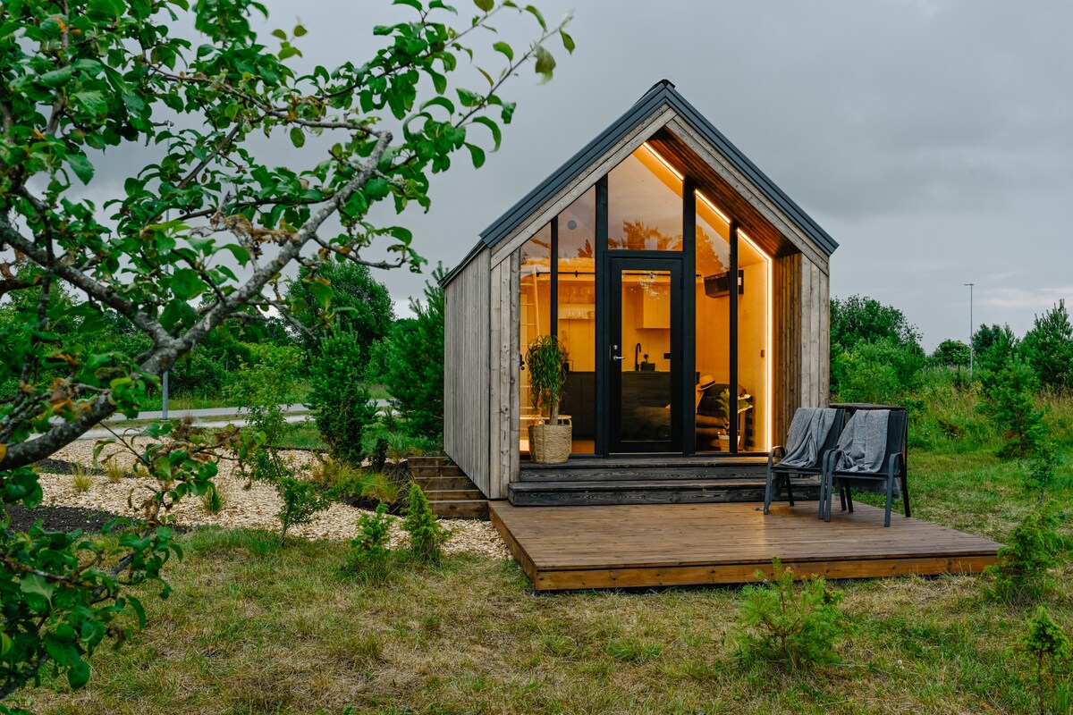 A modern tiny house is showcased, framed by greenery and featuring a wooden deck. Floor-to-ceiling windows illuminate the interior with a warm glow. Two outdoor chairs are positioned on the deck, offering a cozy spot to enjoy the surrounding landscape.