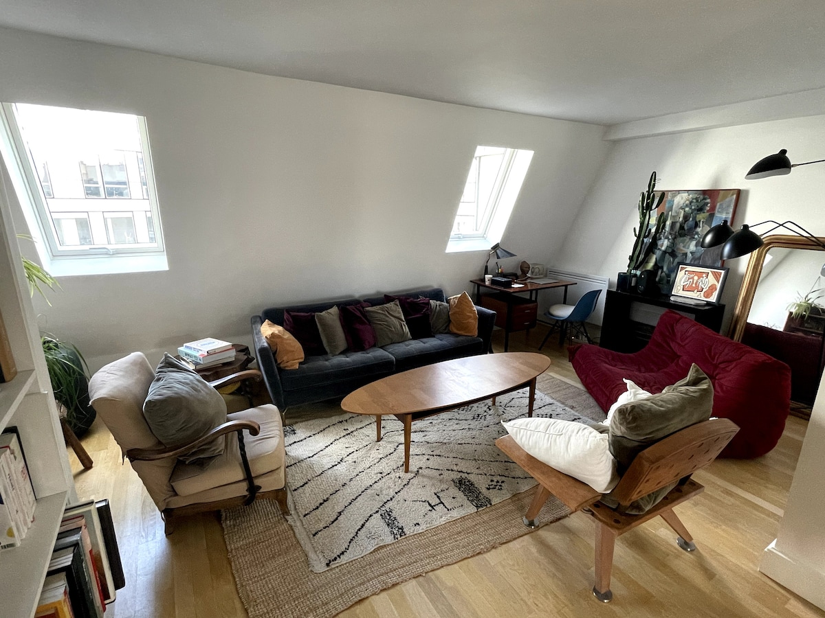 A comfortable living area is furnished with a dark sofa, a red chair, and a wooden coffee table centered on a patterned rug. Natural light enters through two triangular windows, highlighting the eclectic decor and plants that enhance the cozy ambiance.