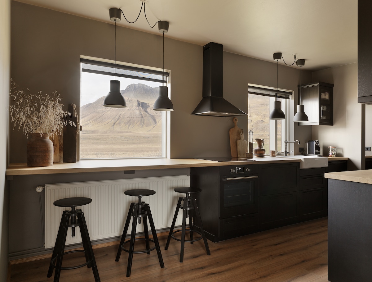 A modern kitchen is illuminated by natural light from dual windows framing mountain views. A minimalist design features dark cabinets, a spacious counter, and three tall stools positioned beside the counter. Black pendant lights hang overhead, adding to the sleek aesthetic.