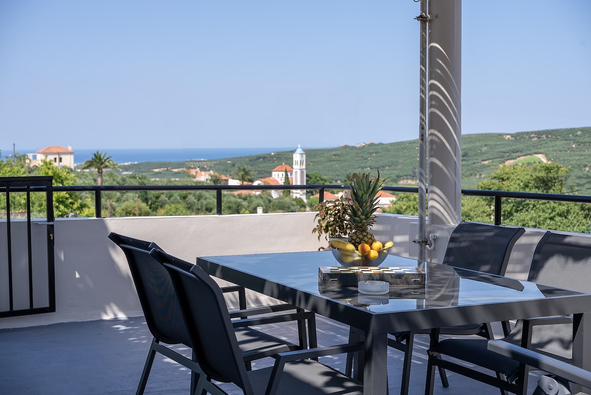 A dining area is visible on the terrace, featuring a sleek table and comfortable chairs. A bowl of lemons is placed at the center of the table. The background showcases a panoramic view of the sea and surrounding landscape, accented by nearby buildings and greenery.