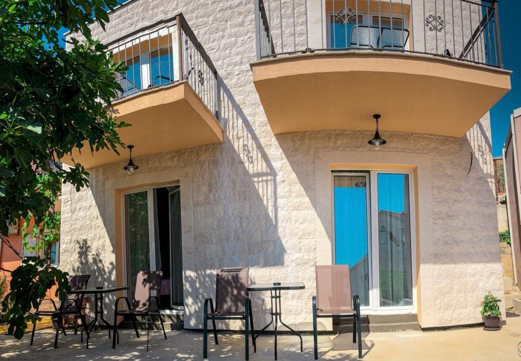 The exterior of the newly constructed apartment building features two balconies with black railing and outdoor seating arrangements. Large glass doors provide access to each unit, allowing natural light to enter. Lush greenery partially obscures the view, contributing to the tranquil setting.