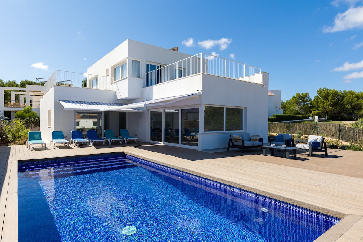 A modern villa features a vibrant pool surrounded by a wooden deck with sun loungers and seating areas. Large glass doors open to a spacious interior, allowing natural light to fill the area. Lush greenery is visible in the background, enhancing the outdoor setting.