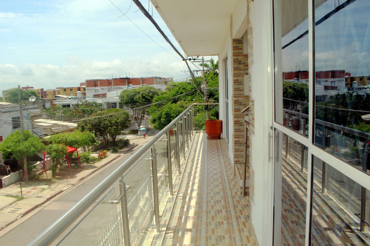 A long balcony extends along the side of the building, featuring glass railings that provide an unobstructed view of the street below. Potted plants add a touch of greenery, while the surrounding area presents a mix of residential and commercial buildings under a clear sky.