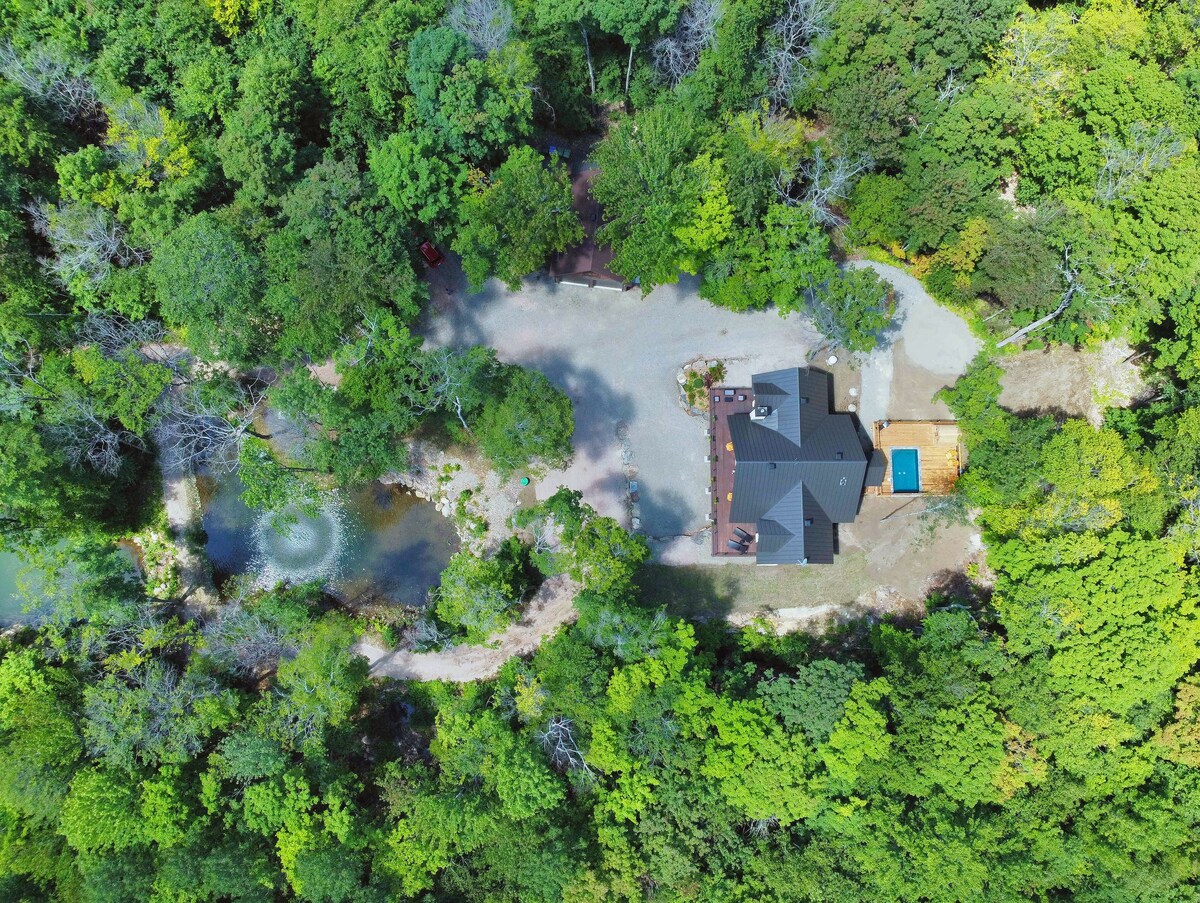 An aerial view presents the chalet surrounded by lush greenery, with a pool visible in the backyard. A winding pathway leads to the property, which is nestled among trees, creating a tranquil and secluded setting. A small pond can be seen nearby.
