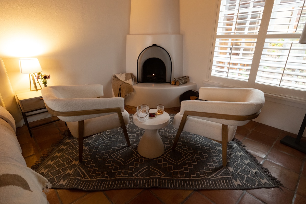 A cozy seating area features two cream-colored chairs positioned around a small, round table. A wood-burning fireplace is situated nearby, and the space is adorned with a decorative rug. Soft lighting from a lamp casts a warm glow on the surrounding walls.