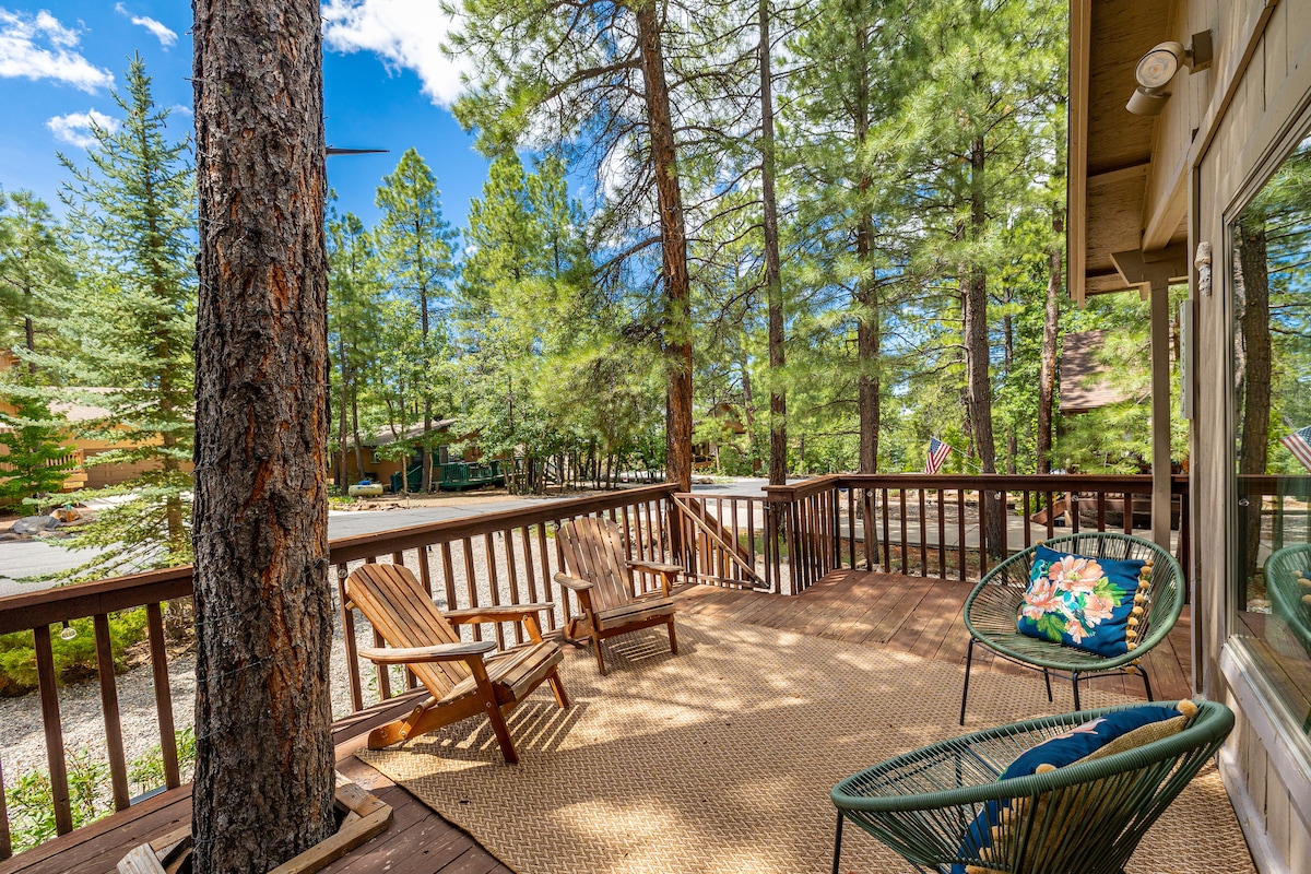 A spacious deck is surrounded by tall pine trees, offering a serene space for relaxation. Two wooden lounge chairs are positioned on a natural rug. A decorative cushion adds a touch of colour to one of the chairs. The area is well-lit by natural sunlight.