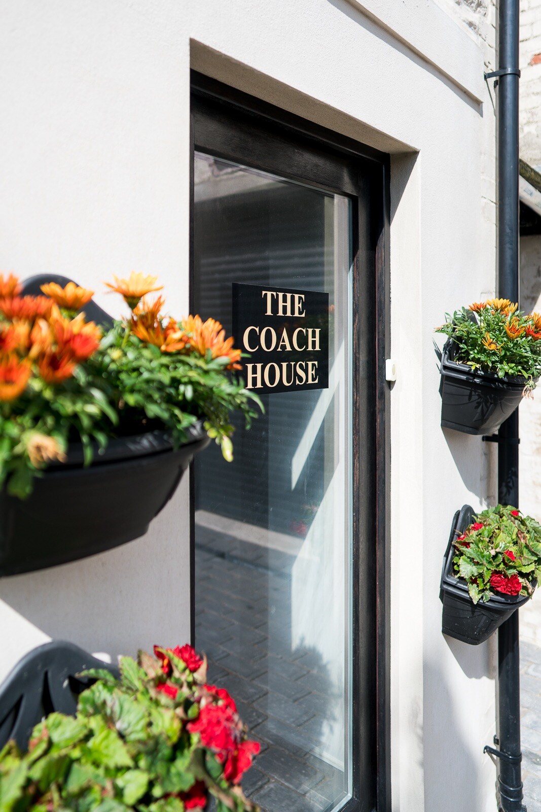The entrance to The Coach House is framed by planters containing vibrant flowers in black pots. A sign with the name 'THE COACH HOUSE' is displayed on the door, offering a welcoming touch to the entranceway.