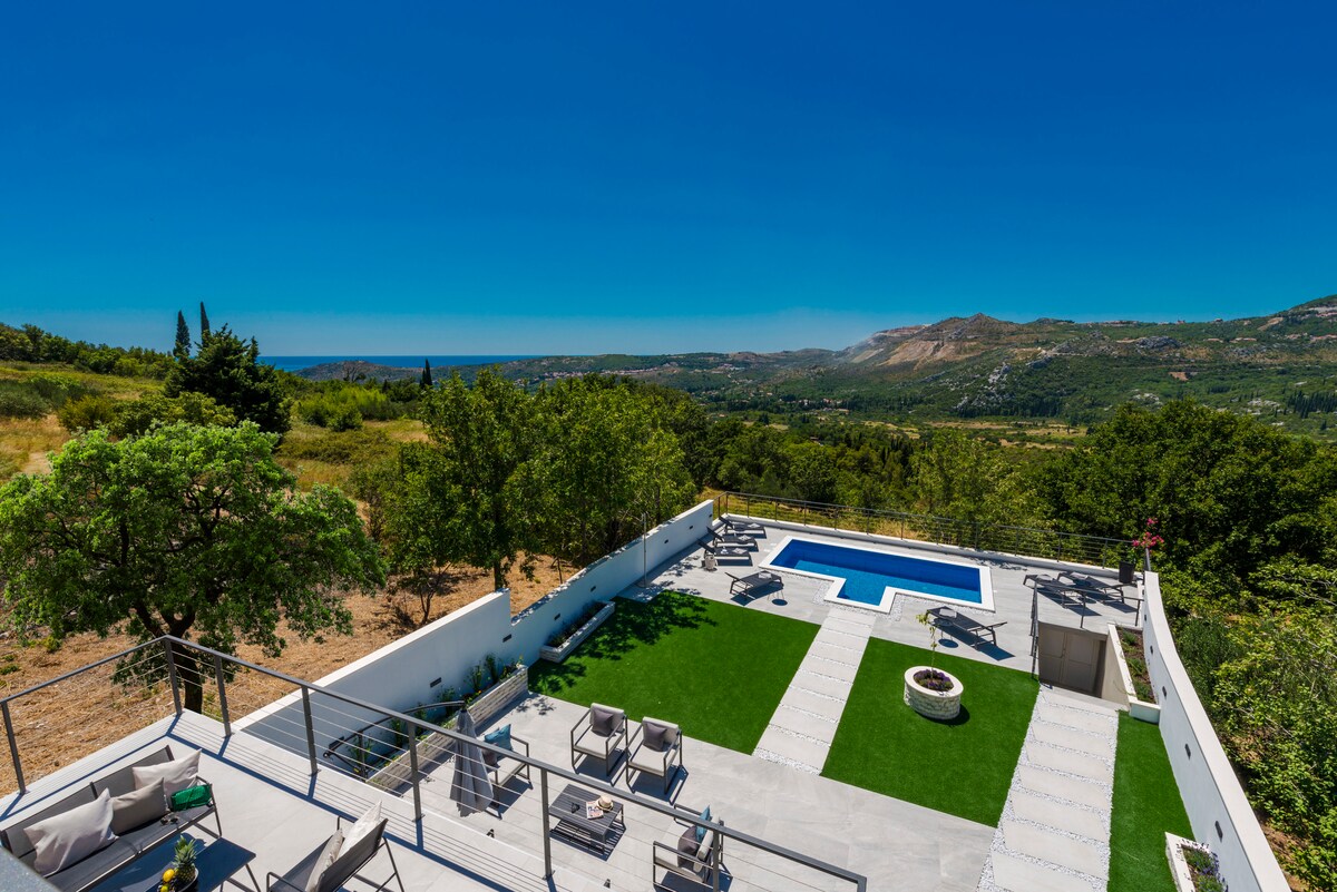 An expansive outdoor area features a private pool surrounded by sun loungers and green grass. The serene landscape reveals lush hills and clear blue skies in the distance, offering a peaceful retreat for relaxation and leisure.
