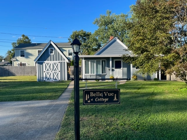 A welcoming view of Belle Vue Cottage features a well-kept lawn with a sign indicating its name. The cottage is framed by trees, with a driveway leading to it. A garage is visible in the background, contributing to the serene atmosphere of the neighborhood.