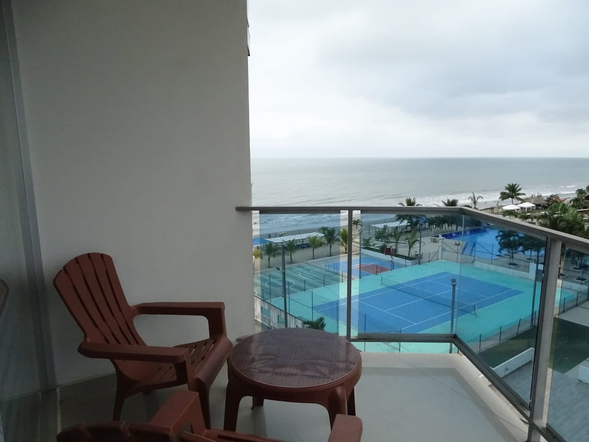 A balcony features two brown chairs and a round table, offering a view of the ocean and nearby tennis courts. The scene is complemented by palm trees and a cloudy sky, creating a serene outdoor space.