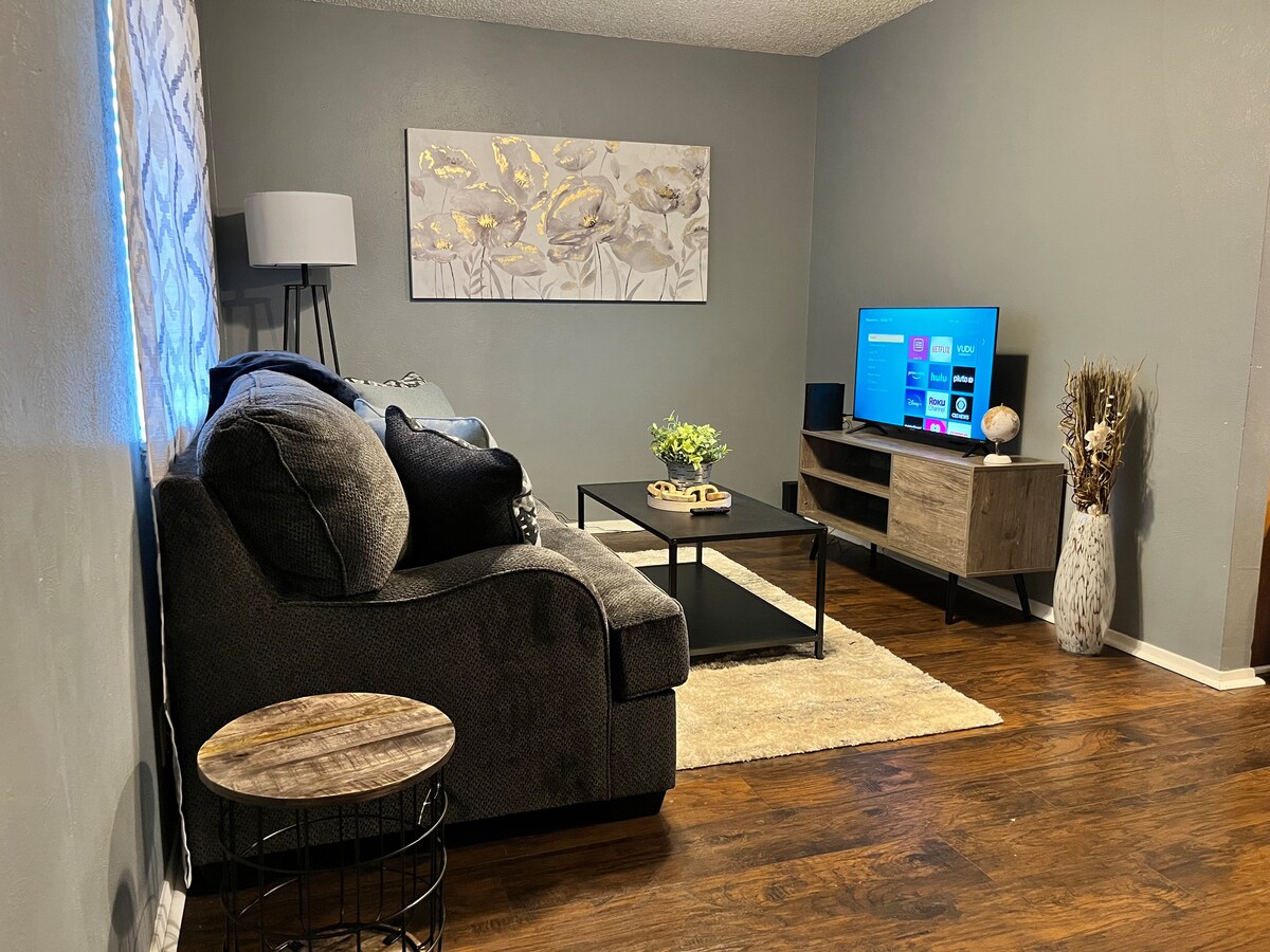 A welcoming living area is presented with a dark sectional sofa, a small round side table, and a rectangular coffee table. A flat-screen TV is mounted on a light wood console, while decorative elements, including a potted plant and artwork, provide a pleasing ambiance.