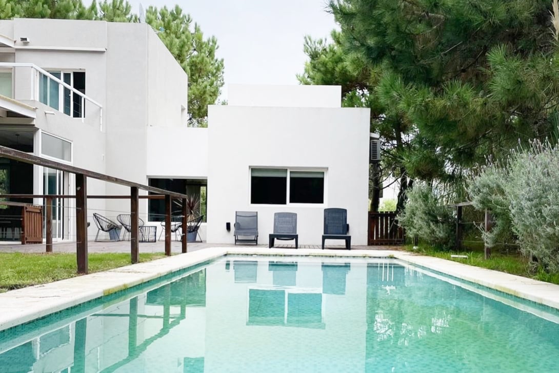 A serene outdoor space features a crystal-clear swimming pool reflecting the surrounding greenery. Four loungers are positioned along the poolside, inviting relaxation. The modern house is visible in the background, with large windows allowing natural light to enter the interior.