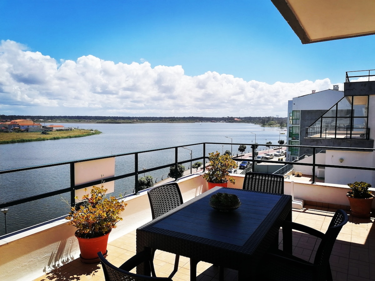 An outdoor terrace features a rectangular dining table surrounded by four chairs. Potted plants add greenery on either side. The expansive view showcases calm waters beneath a clear blue sky, with light clouds creating a serene backdrop.