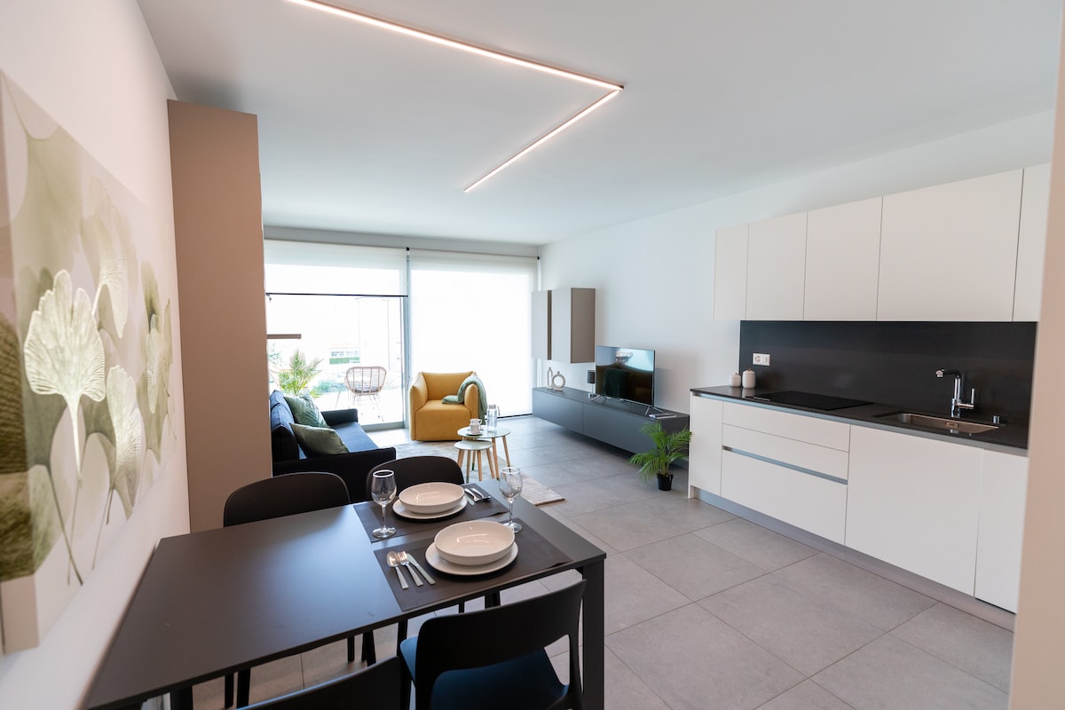 An open living space showcases a modern kitchen with sleek cabinetry and a dining area. A large window allows natural light to illuminate the room, featuring a cozy seating area with a green accent chair and neutral decor, complemented by light tile flooring.