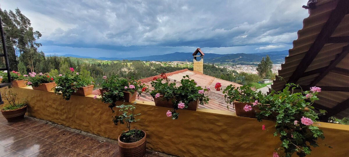 A terrace is adorned with flower pots containing blooming plants, offering a vibrant view of the surrounding landscape. The distant mountains and valleys are visible under a cloudy sky, while the tiled roof and eaves of the house frame the scene.