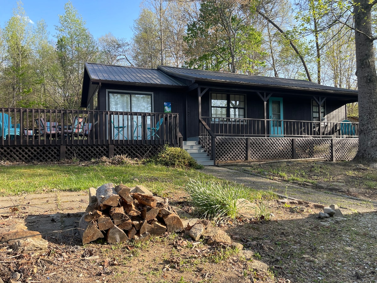 A cozy cabin is set amidst lush greenery, featuring a spacious wooden deck and a distinctive blue door. A stack of firewood is positioned in the foreground, with a fire pit visible nearby. The cabin is surrounded by trees, creating a serene atmosphere.