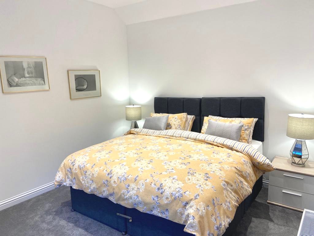 The spacious master bedroom features a large super-king bed dressed in a floral yellow duvet cover. Bedside tables with lamps are positioned on either side, complemented by framed artwork on the walls. Natural light brightens the neutral-colored room, enhancing the overall serenity.