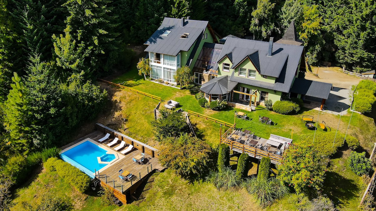 An aerial view captures two houses nestled among lush trees. A wooden deck surrounds a rectangular pool, with lounge chairs arranged neatly nearby. The well-kept grounds feature areas for relaxation, highlighted by natural greenery and pathways leading towards the property.
