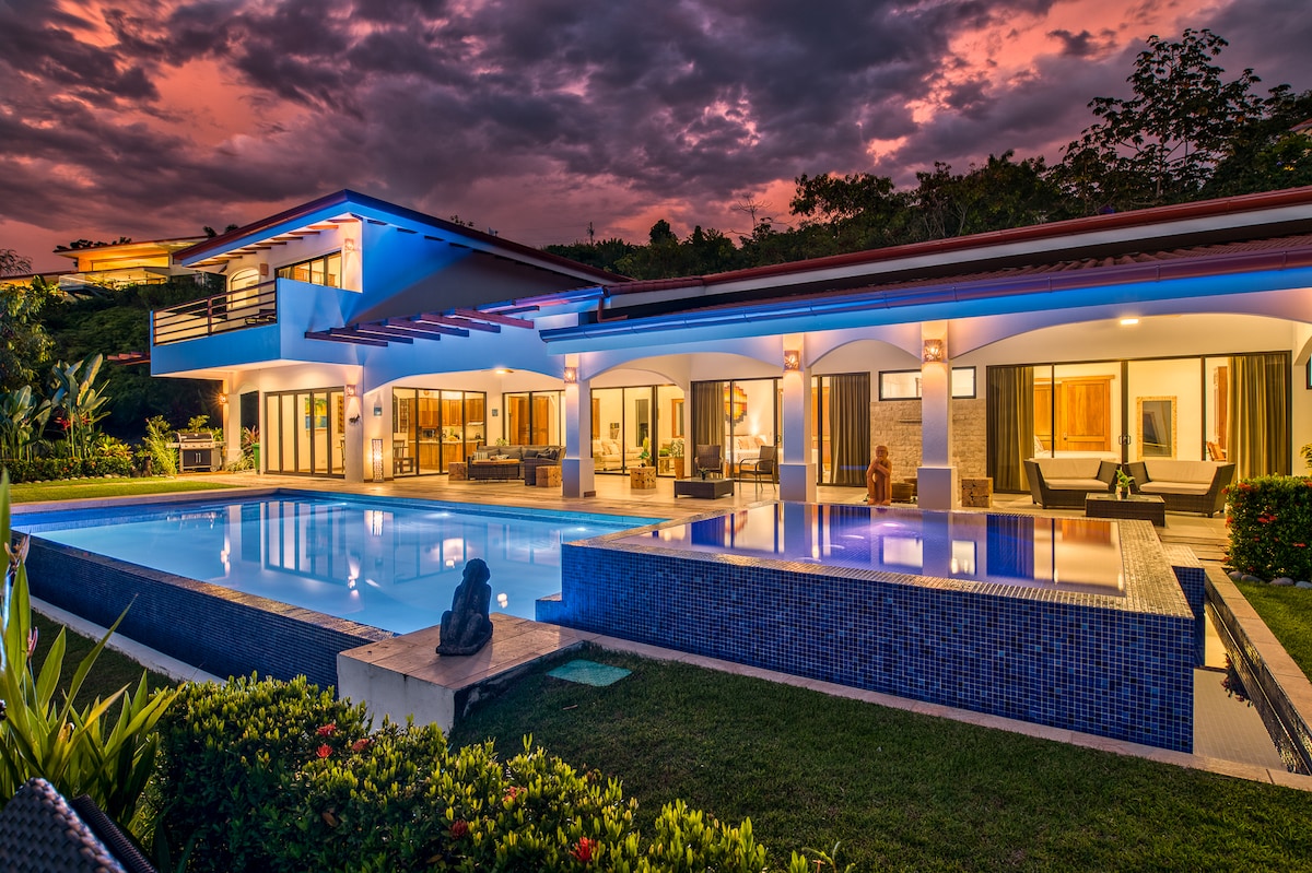 The villa features a modern design with a sprawling layout, illuminated by evening lights. An infinity pool reflects the vibrant sunset sky, while comfortable seating areas are positioned nearby, inviting relaxation amidst the lush gardens.