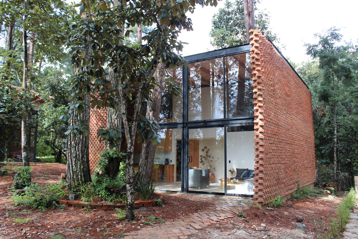 A contemporary loft is surrounded by lush greenery, showcasing a blend of textured brick and expansive glass walls. The unique architectural design invites natural light, creating an open and inviting exterior that harmonizes with the forested setting.