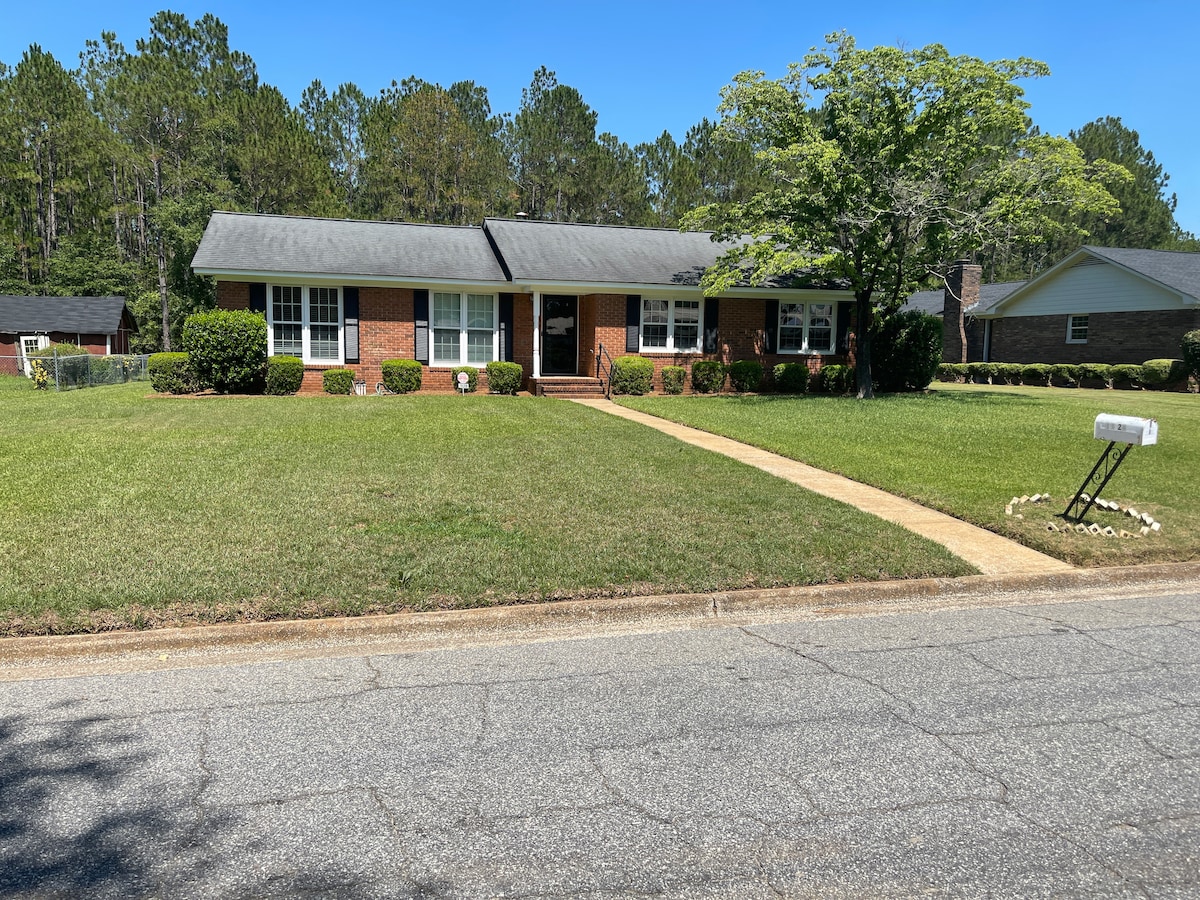 A charming single-story brick home is set amidst a green lawn, bordered by well-maintained hedges. The front walkway leads to a covered porch, flanked by two large windows. Surrounding trees provide natural shade, contributing to the peaceful neighborhood setting.