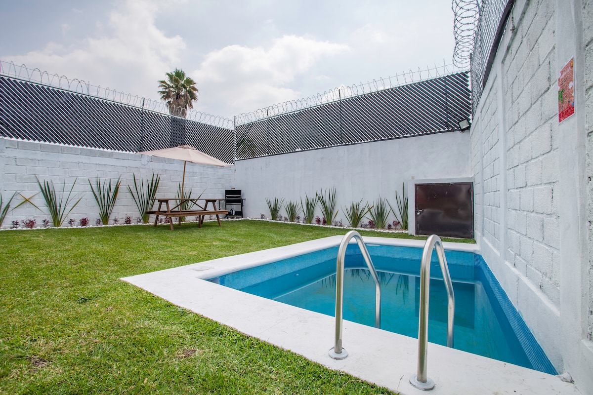 A private pool is set within a well-maintained backyard, featuring manicured grass and neatly arranged plants along the perimeter. A shaded dining area is visible, accompanied by a barbecue. A tall fence provides privacy, with decorative elements enhancing the space.