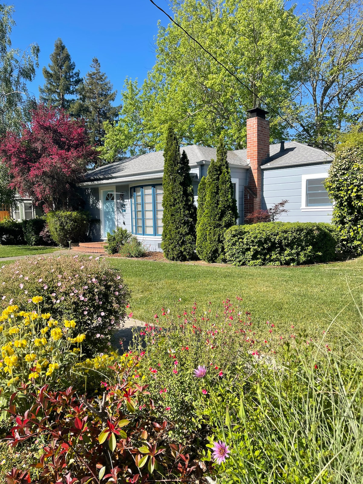 A charming 1940's bungalow is set on a corner lot, surrounded by lush greenery and vibrant flowers. Tall trees provide shade, while a brick chimney adds character to the exterior. The home's blue siding and large front windows create an inviting facade.