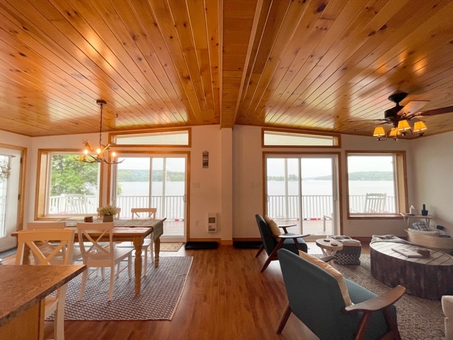 An open living space features a wood-paneled ceiling and large windows offering views of the lake. A dining table with white chairs is positioned near the windows. Comfortable seating, including blue armchairs and a circular coffee table, creates a welcoming atmosphere.
