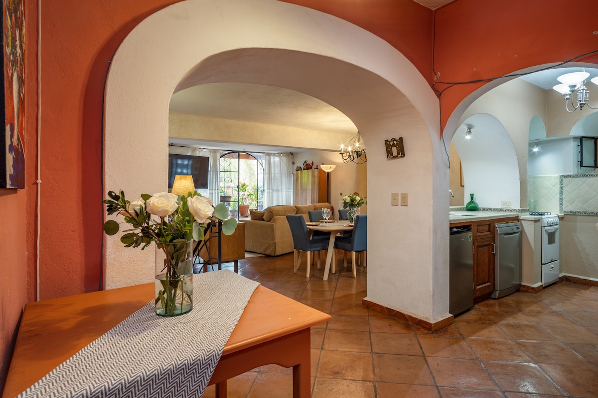 An open living space is captured, showcasing arched doorways leading to various areas. A wooden table is set with a vase of white roses, while a cozy sofa and dining table with colorful chairs are visible. The kitchen area features modern appliances.
