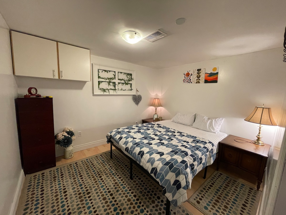 A bedroom features a spacious bed with a patterned quilt and matching pillows, flanked by two bedside lamps. A small dresser and wall art add character to the room. Soft lighting creates a cozy environment, complemented by a subtle area rug beneath the bed.