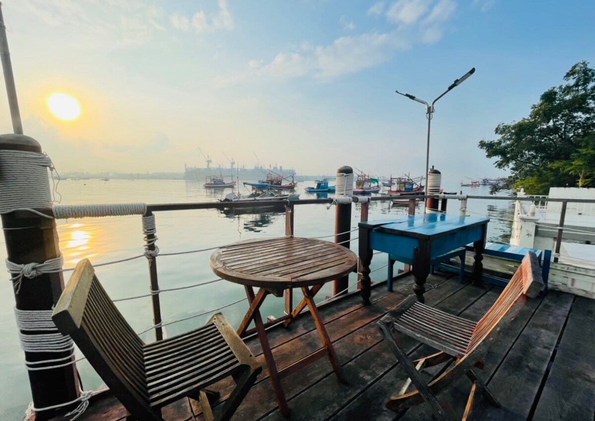 A wooden deck overlooks the calm waters, featuring a round table and two chairs. In the background, boats are anchored, with a soft sunrise illuminating the scene. The tranquil environment is enhanced by the reflections on the water's surface.