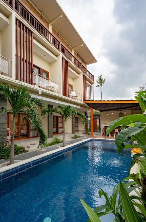 An outdoor swimming pool is framed by lush greenery and palm trees, inviting relaxation. The two-story guesthouse features a balcony on the upper level with wooden railings, and large windows allow natural light to fill the interior spaces.