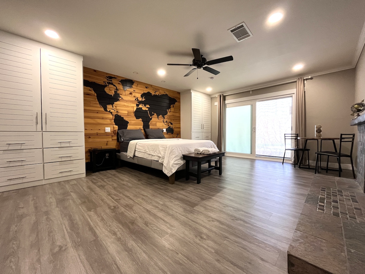 A spacious guest room features a world map accent wall, complemented by light wood paneling. A large bed is centered on modern flooring, with a seating area including a small table and two chairs. Natural light streams through patio doors leading outside.