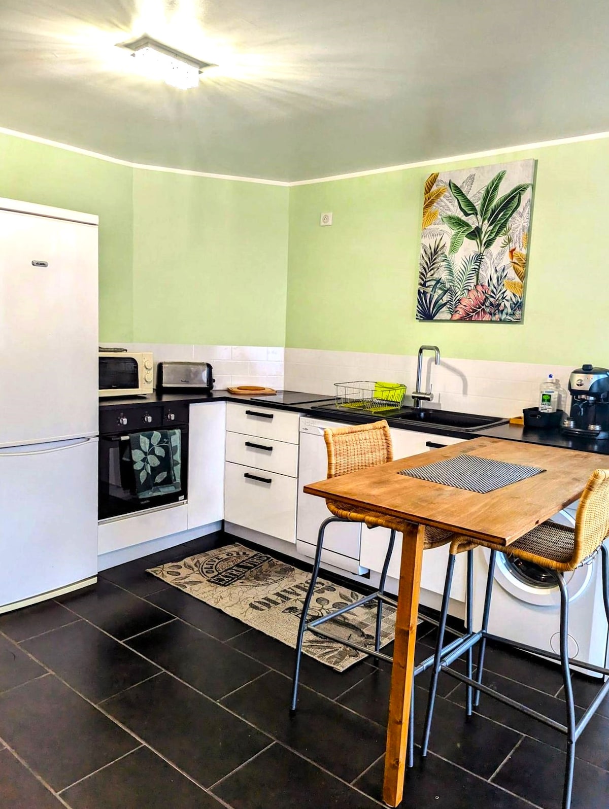 The kitchen features modern appliances, including a refrigerator, oven, and microwave. A wooden dining table with stools complements the space, which is accented by a green wall and a tropical-themed wall art piece. Natural light brightens the area, enhancing its functionality.