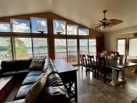The open concept living space features large windows that offer expansive views of the lake, allowing natural light to fill the area. A dark leather sectional sofa is positioned near a dining table, enhancing the inviting atmosphere, while a ceiling fan provides comfort.