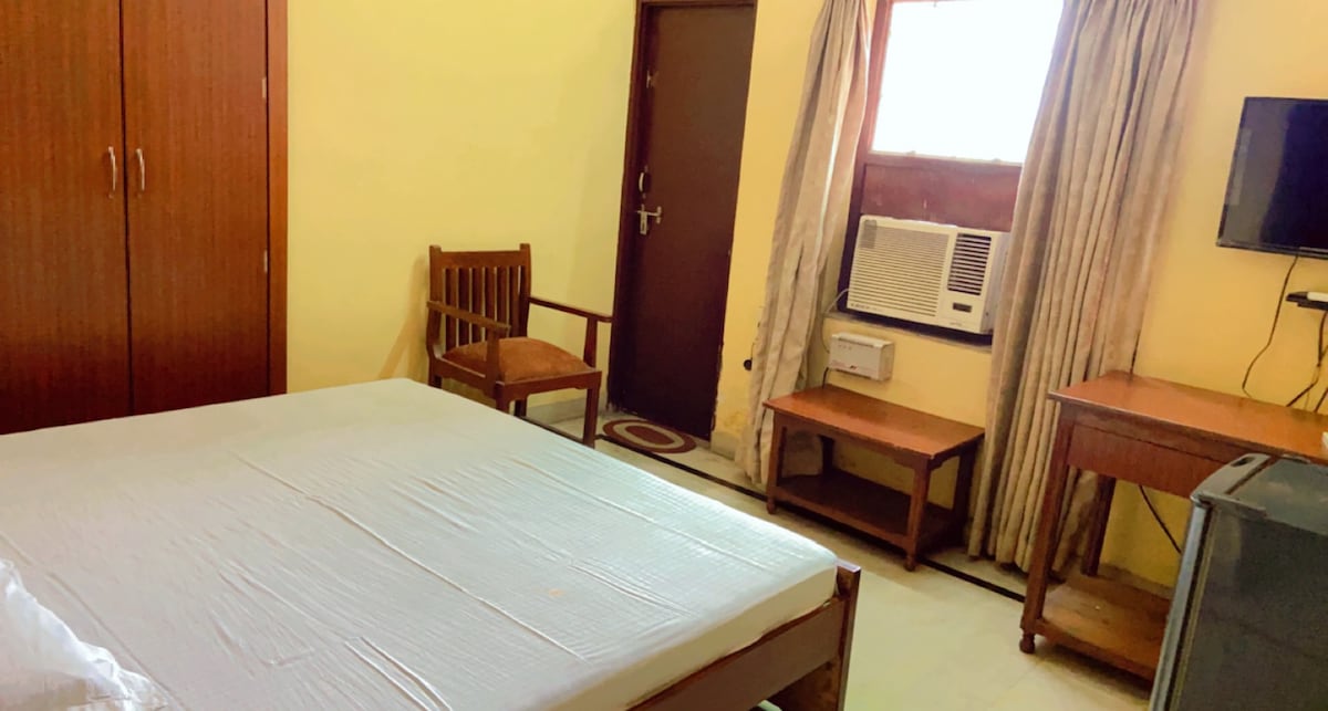 A simple bedroom features a spacious bed with a plain mattress. An air conditioning unit is mounted above a small desk and a mini refrigerator. A wooden chair is positioned near the bed, and curtains frame a window, allowing natural light to enter.