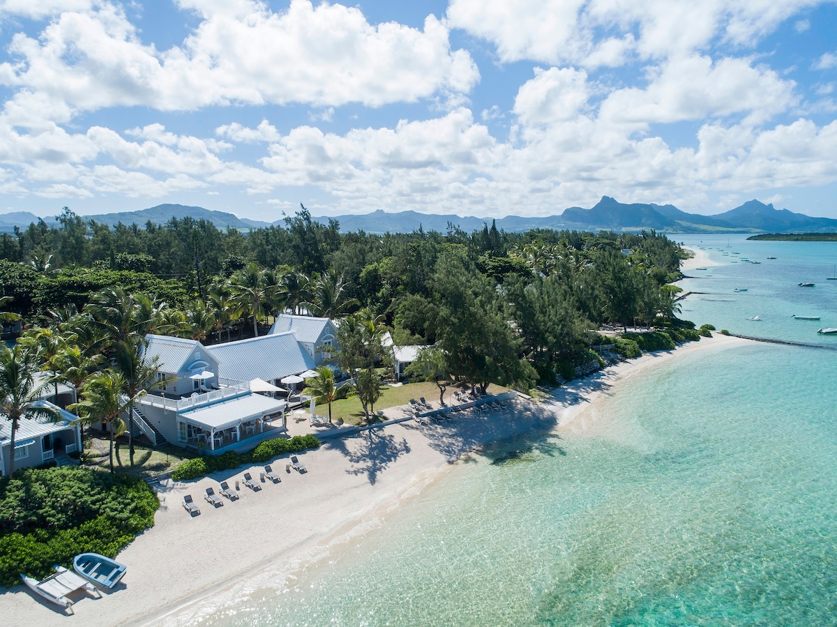 An aerial view showcases the boutique hotel near a white sandy beach and turquoise lagoon, surrounded by lush greenery and palm trees. Sun loungers are positioned along the shoreline, with boats gently floating in the water against a backdrop of distant mountains and blue skies.