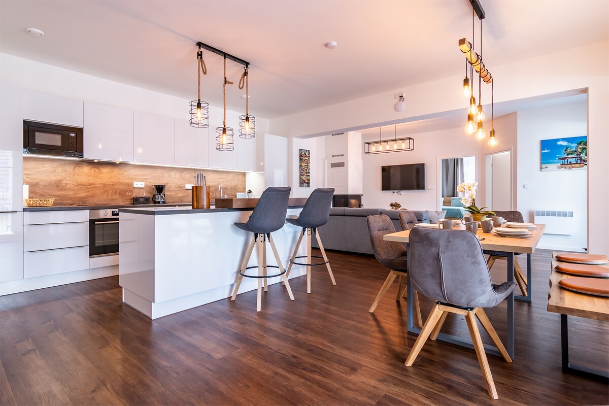 A modern open-concept living area features a sleek kitchen with white cabinetry and a wooden backsplash. Bar stools are positioned at a kitchen island, while a dining table with grey upholstered chairs is set nearby. Soft lighting fixtures enhance the inviting space.