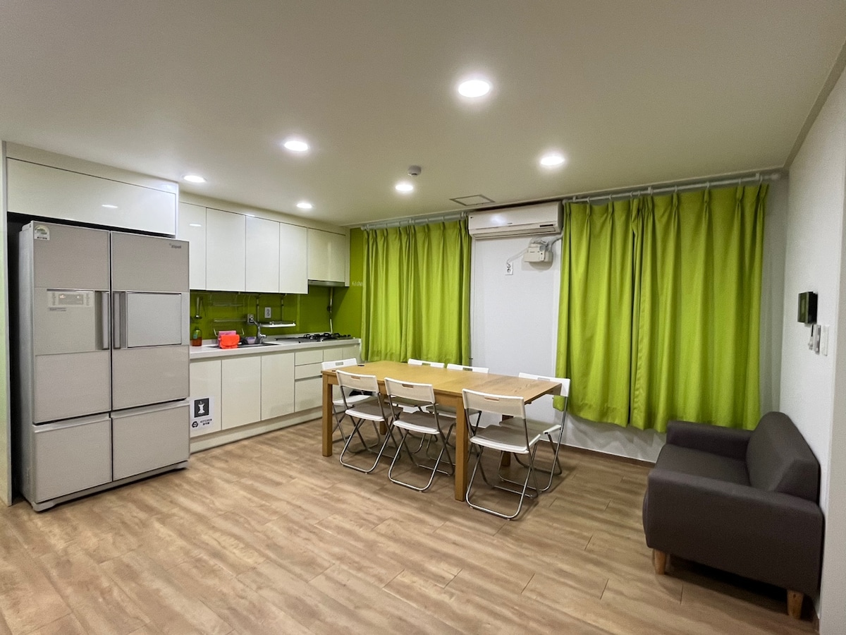 A bright kitchen area is complemented by sleek, modern appliances, including a refrigerator. A dining table is set with several white chairs, all enhanced by vibrant green curtains that frame the windows. A cozy brown sofa is positioned in the corner, providing additional seating.