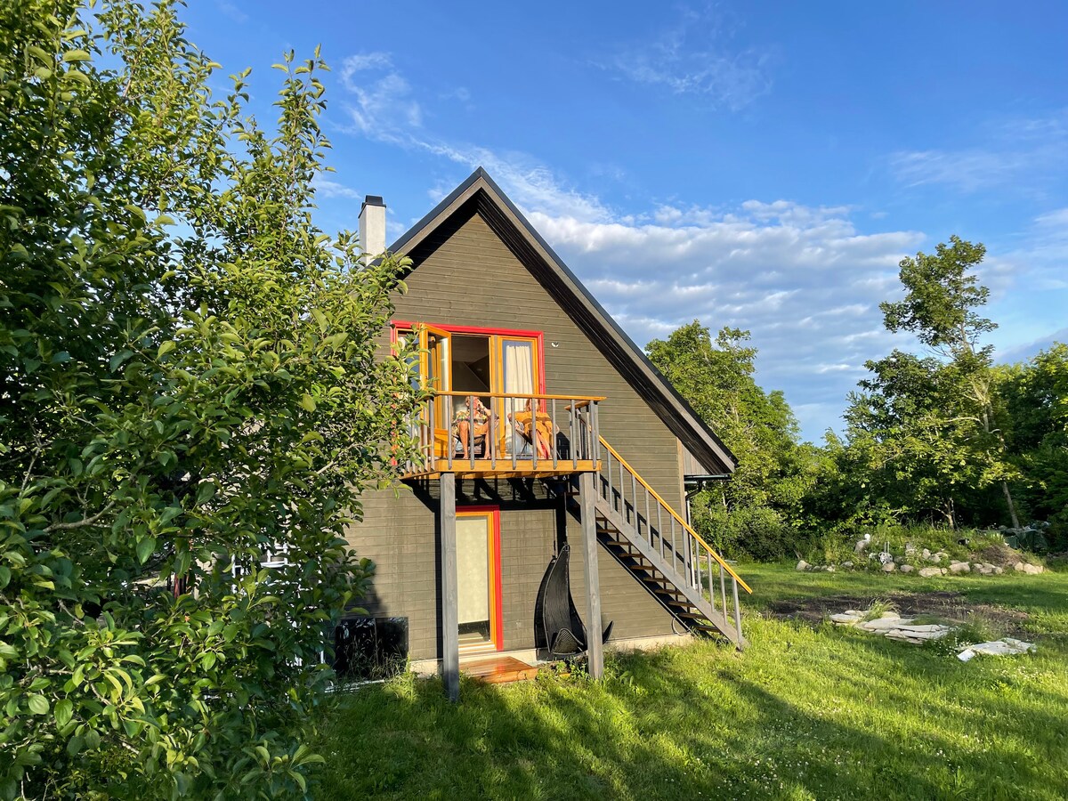 A modern two-story building with a wooden deck and bright orange accents is surrounded by green trees and grass. A staircase leads to the upper level, where guests can enjoy the outdoors from the deck, evident in the inviting setting.