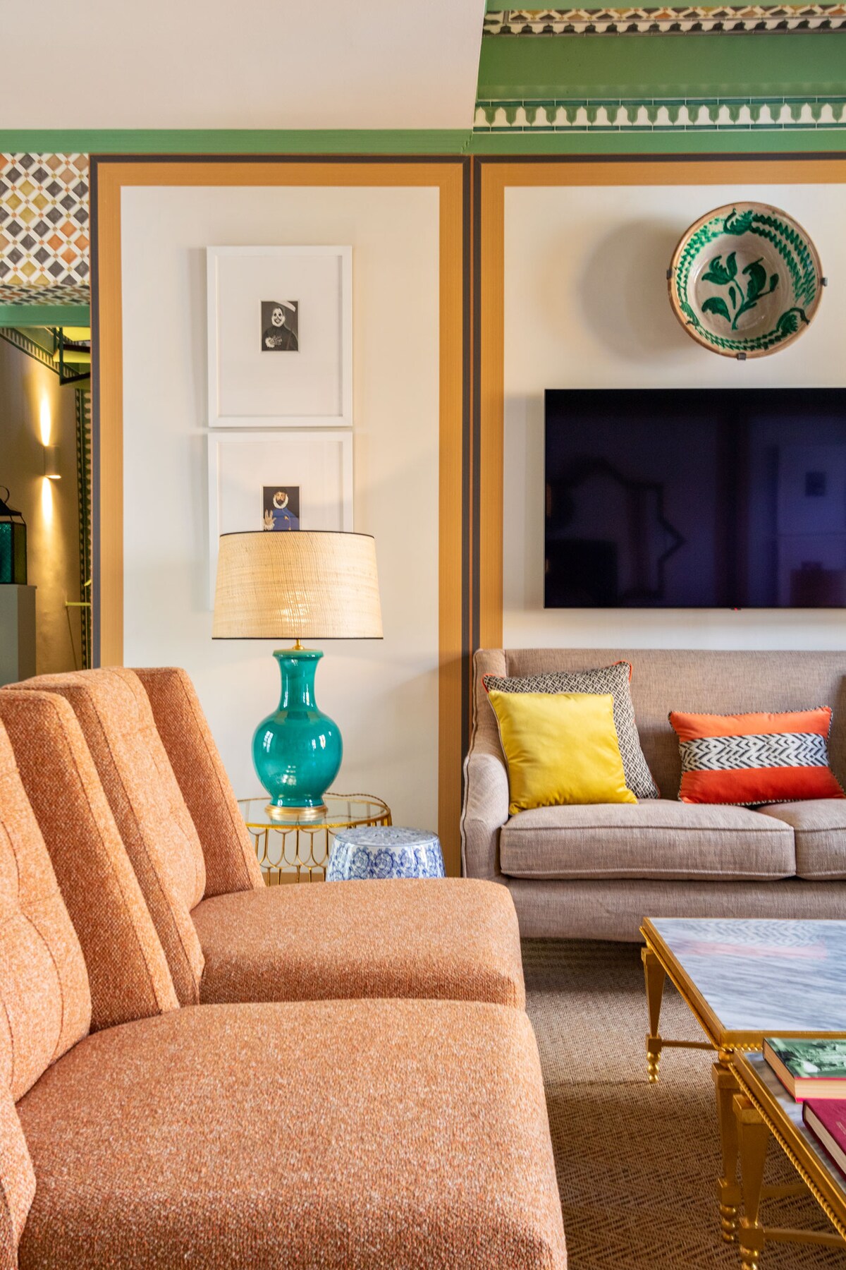 A spacious living area is presented with two comfortable couches in warm tones. A turquoise lamp stands on a side table, complementing colorful cushions on the sofa. A flat-screen television is mounted on the wall, contributing to a modern decor.