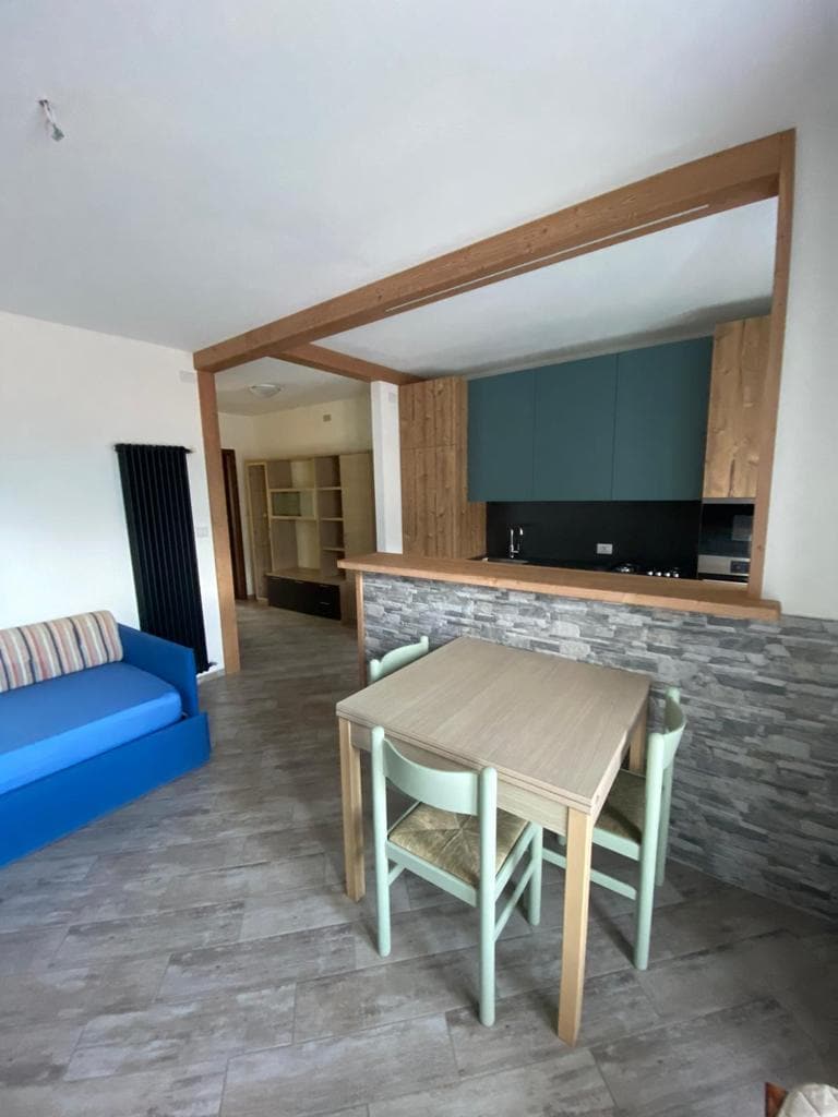 A modern living area features a blue sofa and a light wood dining table surrounded by four pastel green chairs. The adjoining kitchen is visible, characterized by blue cabinetry and stone accent details. Natural light illuminates the open layout, enhancing the sense of space.