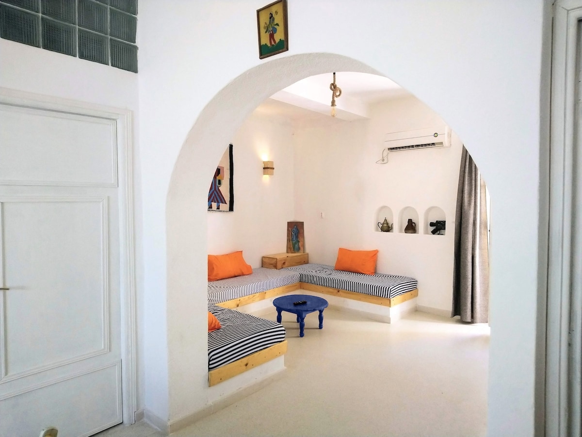 A light-filled living area features two built-in seating areas covered with striped cushions in orange and blue. A small blue table sits at the center, and decorative niches on the wall display various items. An archway enhances the openness of the space.
